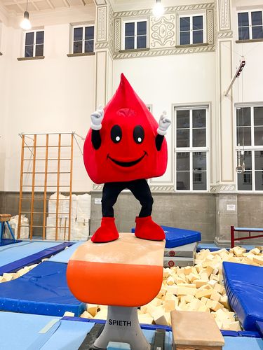 Ein rotes tropfenförmiges Maskottchen macht eine winkende Pose in einer Sporthalle