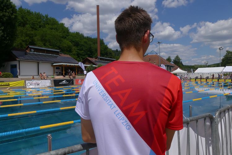 Man sieht den Rücken einer Person, die vor einem Schwimmbecken steht und die Uni Leipzig Wettkampfkleidung trägt.