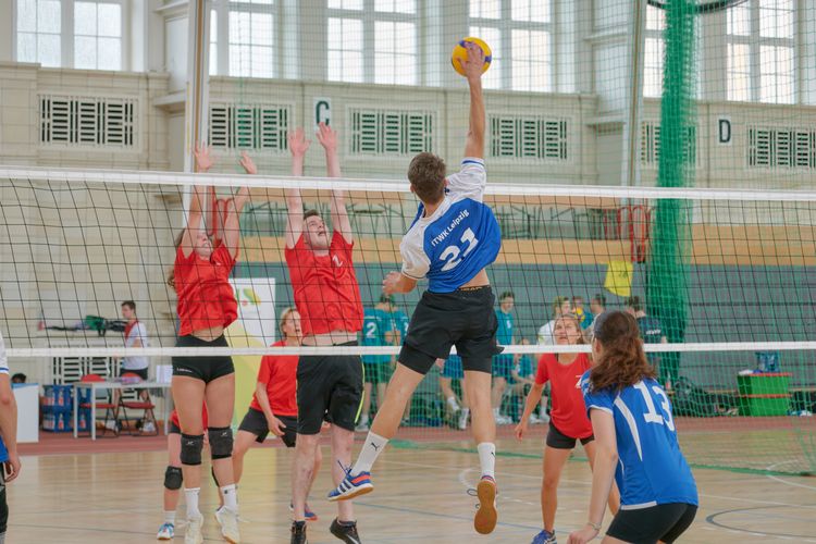 Man sieht eine Nachaufnahme einer Angriffssituation im Volleyball. Drei Spieler:innen befinden sich im Sprung am Netz. Eine Person greift an, die anderen zwei setzen zum Block an.