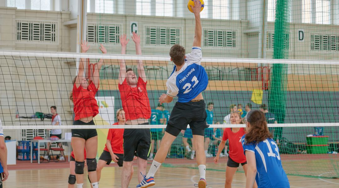 Man sieht eine Nachaufnahme einer Angriffssituation im Volleyball. Drei Spieler:innen befinden sich im Sprung am Netz. Eine Person greift an, die anderen zwei setzen zum Block an.