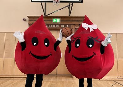 Blutspendeaktion 2024 Zwei Maskottchen halten einen Basketball vor einem Basketballkorb.