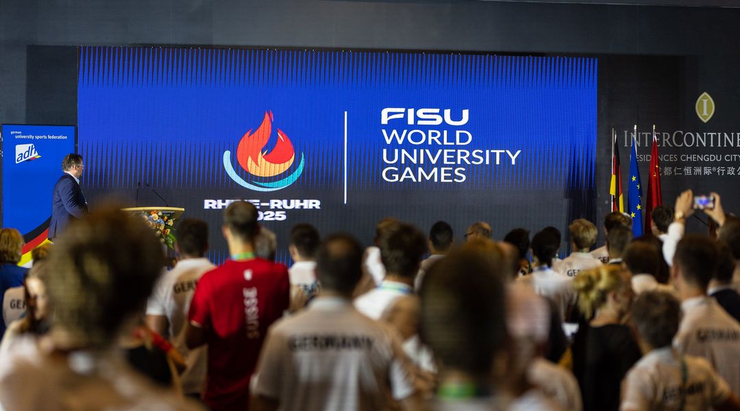 Man sieht eine Anzeigetafel, auf der die neuen FISU Games 2025 angezeigt werden. Verschwommen im Vordergrund sind Rücken und Hinterköpfe von Leuten, die zur Anzeigetafel gerichtet sind.