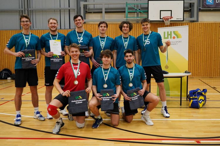 Man sieht eine Gruppe junger Menschen, die in Sportkleidung in einer Turnhalle stehen. Es wird ein Volleyball und eine Urkunde in die Kamera gehalten